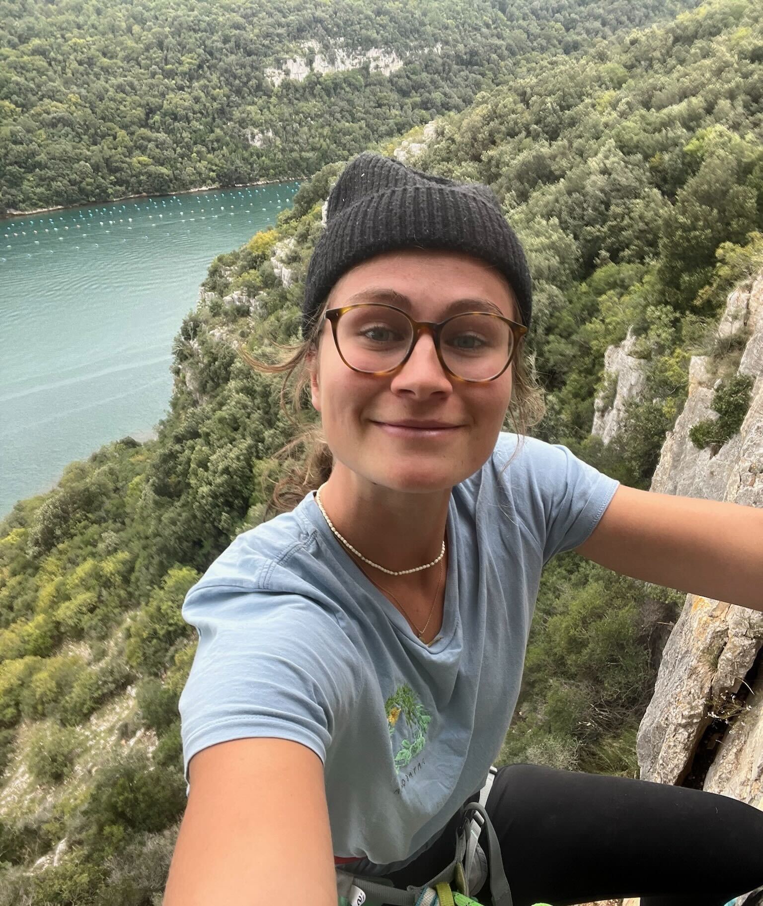 Luiser hängt an einer Felswand im Gurt und macht ein Selfie. Im Hintergrund ein See umrundet mit Bäumen
