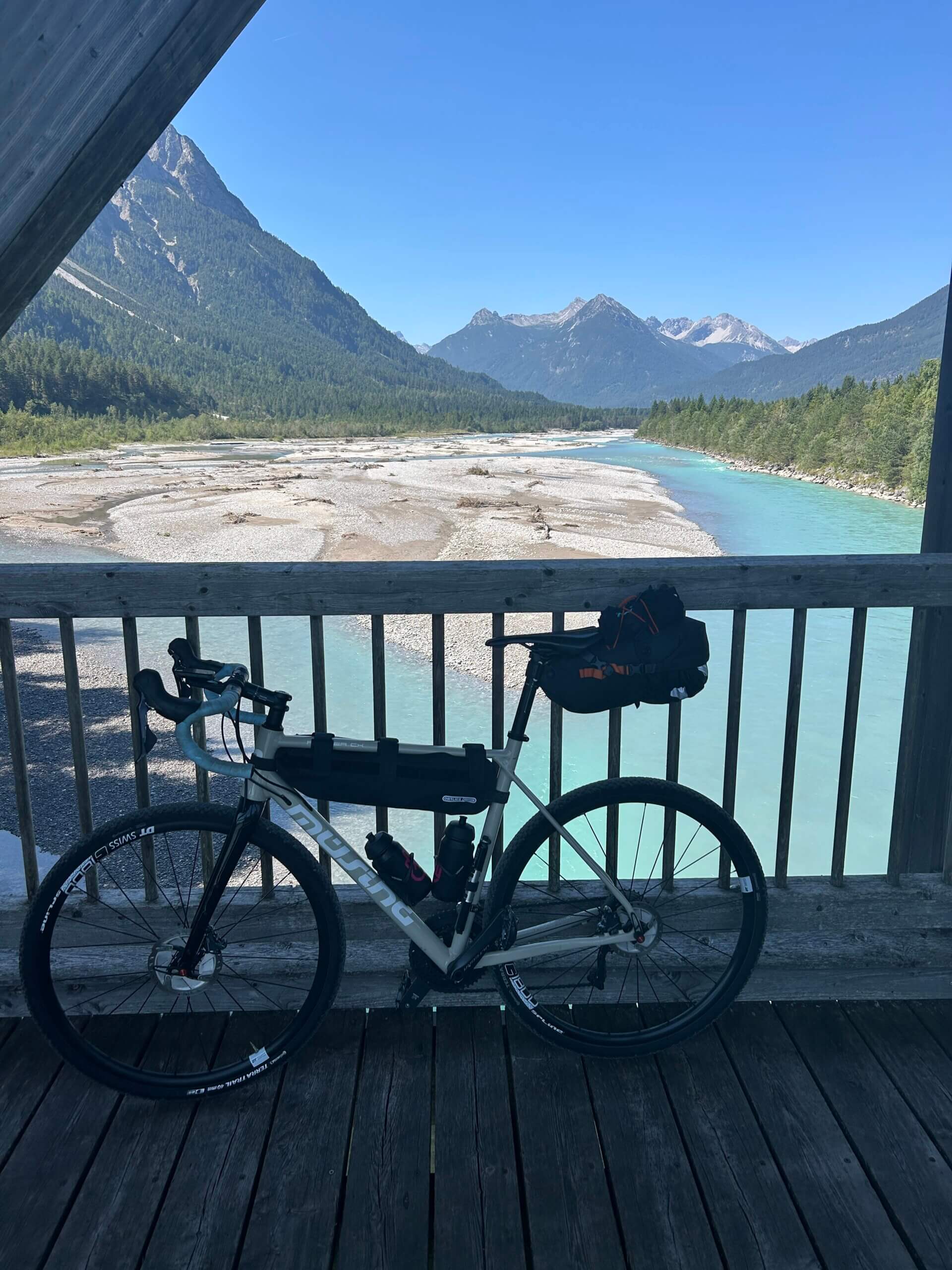 Fahrrad mit Taschen lehnt an Geländer einer Brücke über einen Fluss, im Hintergrund Bergkulisse